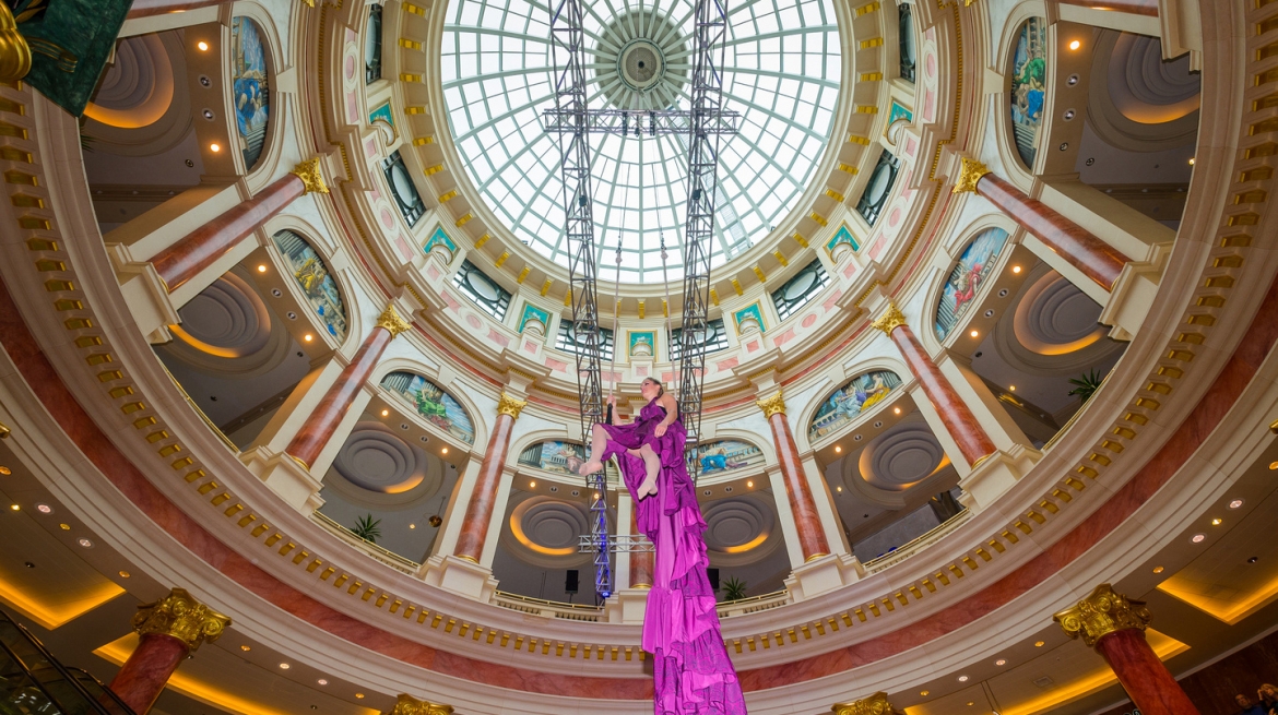 Looking up into the domed roof of a building where a model poses on a trapeze suspended from the glass ceiling. She is wearing a dress with a very long train that trails down.