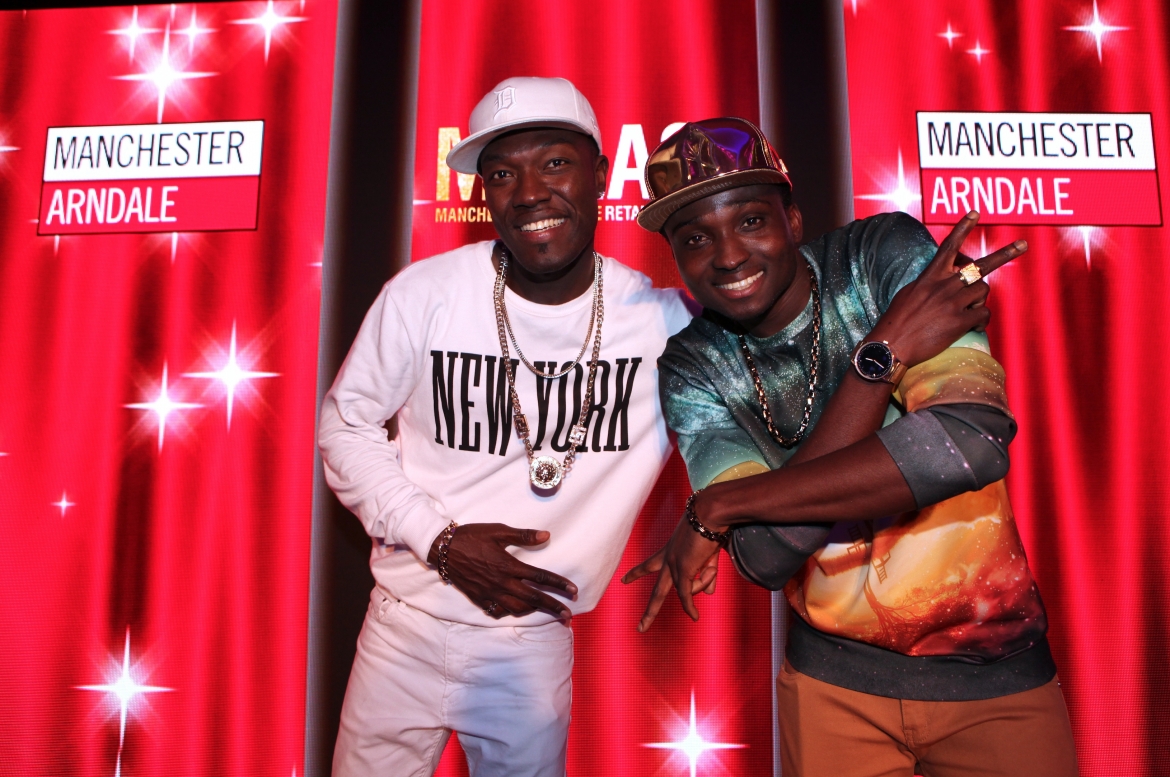 2 young men (Reggie n Bollie), wearing baseball caps and jewellery, pose in front of digital screen at Manchester Arndale Retailer Awards